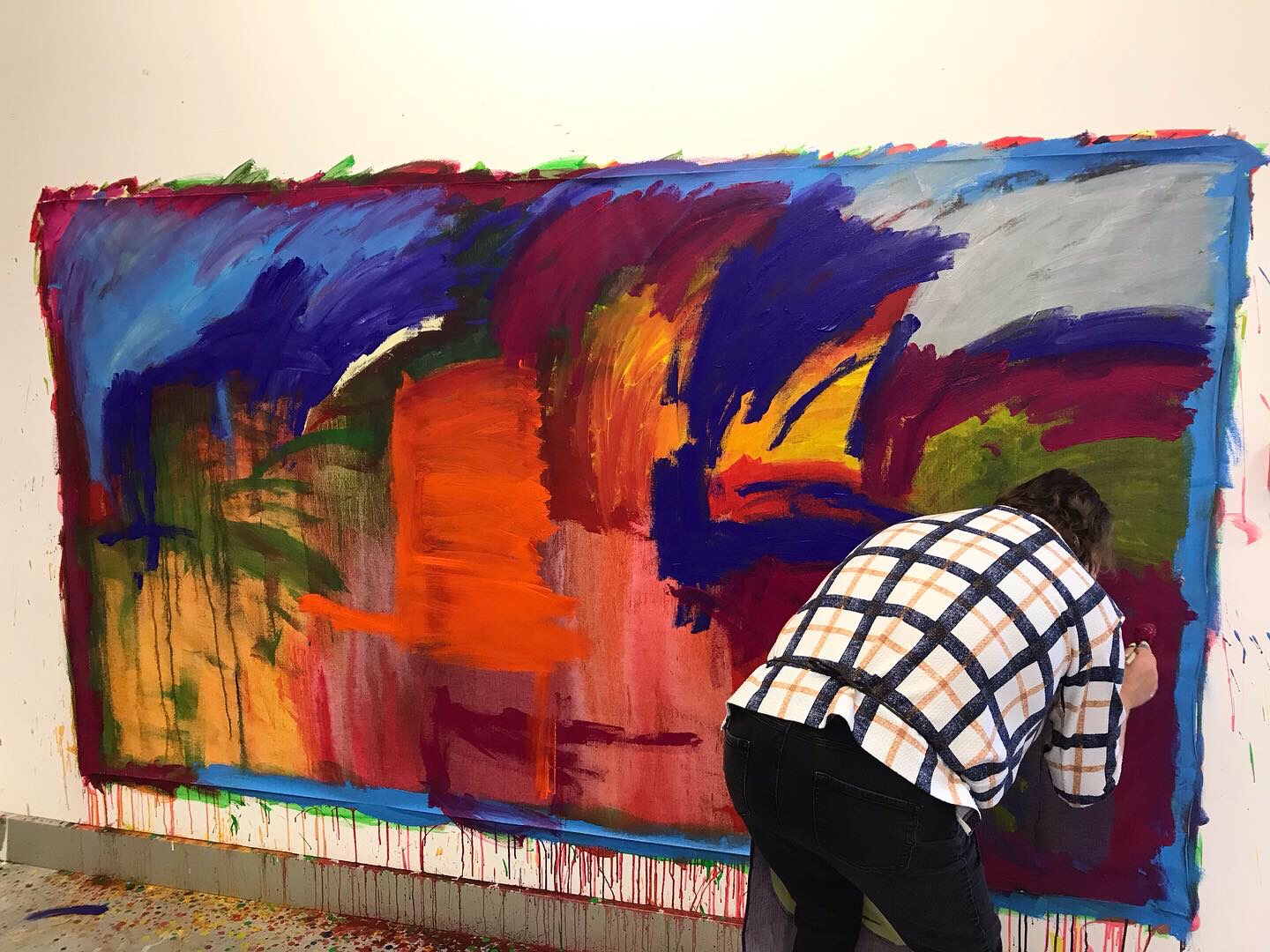 A photo of a person painting a colourful, large abstract painting on the wall. Their back is turned and they are bent over, painting close up to the canvas. 