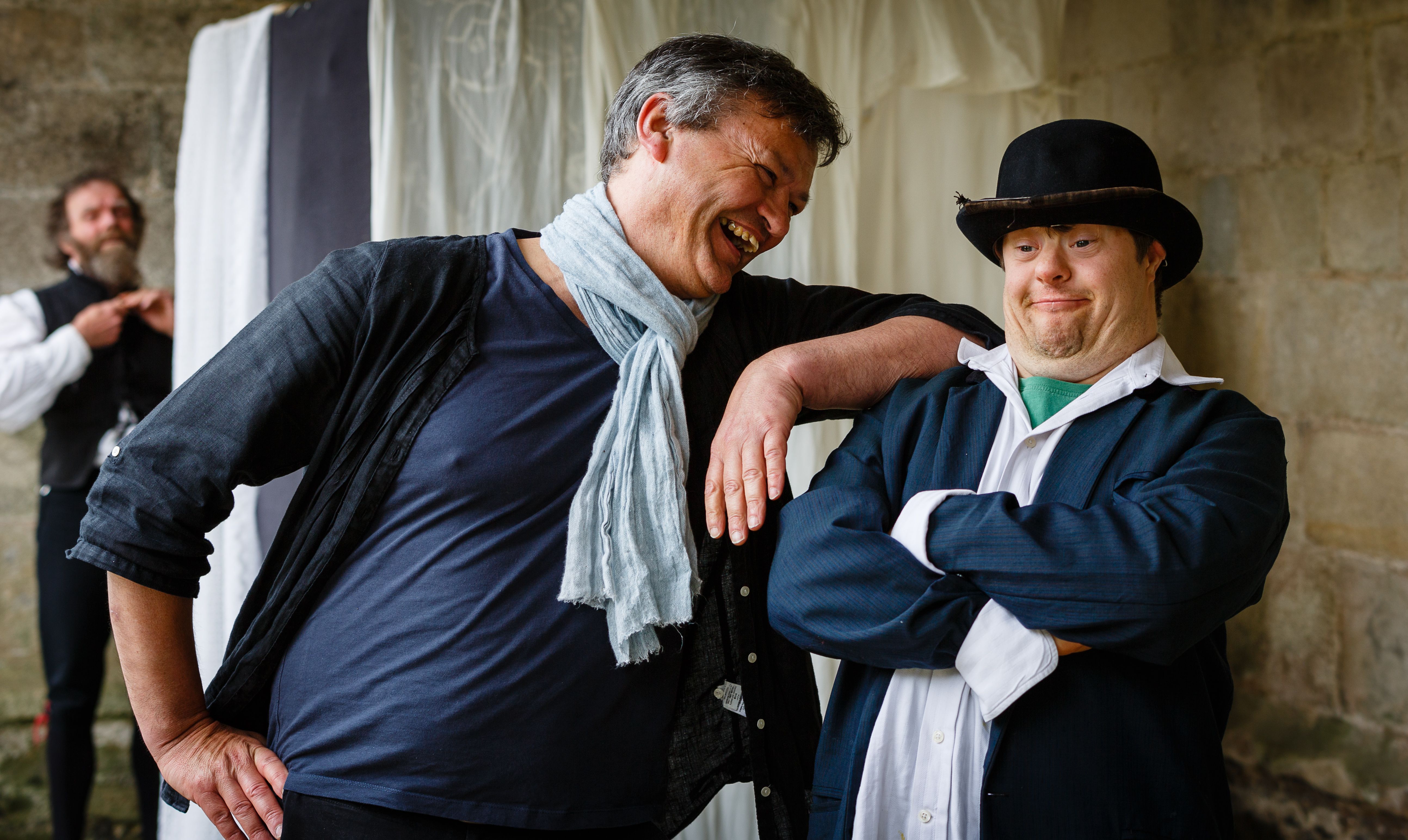 2 people standing next to each other outside a stone building. The person on the left is resting their arm on the other person's shoulder and looking at the other person and smiling. They are wearing a blue tee-shirt and black waistcoat and a light blue scarf. The person on the right is standing with their arms crossed and smiling at the camera. They are wearing a white shirt under a dark blue suit jacket and a black bowler hat. There is a person in the background wearing a white shirt and black waistcoat. 