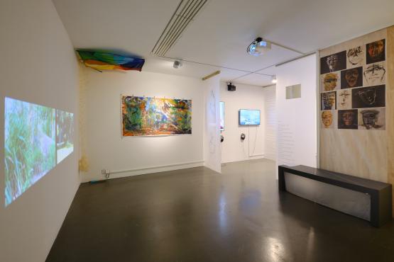film projected on wall, kite on ceiling in corner, large  multi coloured painting, some monitors and words and brown portraits hung above a black benc