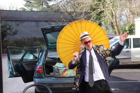 man holding a yellow parasol wearing a tie jacket, shirt and hat with a red nose and sunglasses dancing in front of an open car boot in a car park with a white car and trees in the background