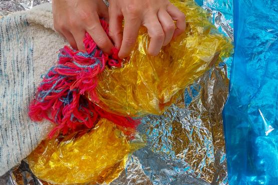 Photograph of hands clutching a wide variety of brightly coloured materials including wool, coloured acetate and woven fabric