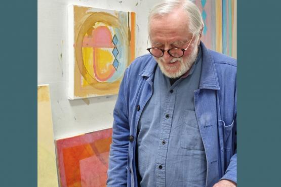 man with grey hair and beard and glasses and blue jacket, looking down in a studio with paintings behind him