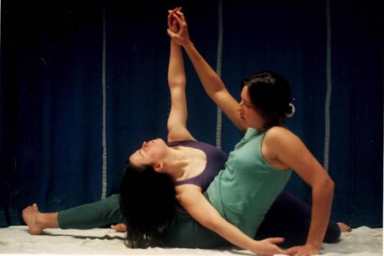 2 people sitting on the floor dancing. They are sitting face to face holding hands with one arm raised upwards. One person on the left is leaning backwards.
