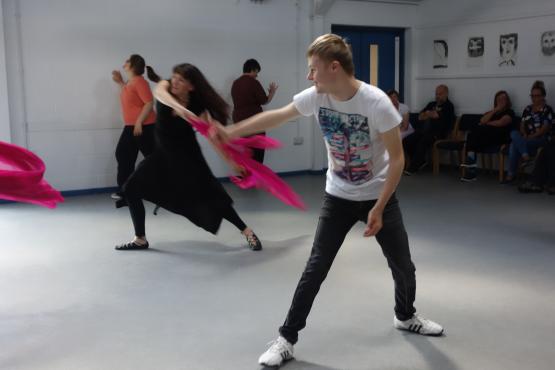 2 people dancing in a hall space. The person in the foreground is reaching one of their arms outwards and smiling. The second person is using a bright pink scarf to dance with. Their are 2 other people in the background dancing.