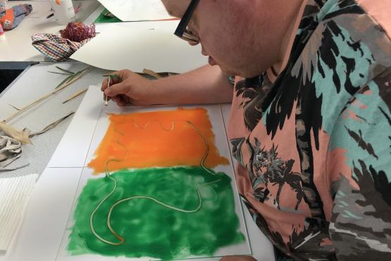 A person sitting at a table monoprinting. They are using orange and green ink, which they have rolled out in 2 block squares. They are using a pencil to draw into the ink. 