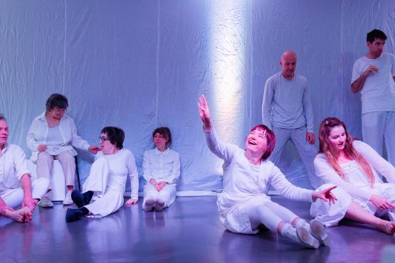 a group of 8 people wearing white in a performance space. 6 people are sitting on the floor resting. 1 person to the right is raising their arm in the air and looking up at it.