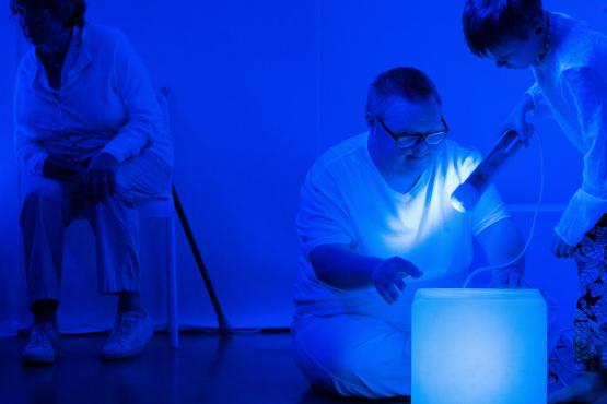 Three people are in a space filled with blue light. An older person sits on a chair. A younger person sits on the floor looking at a large light cube. A child shines a torch light on the floor next to the light cube.
