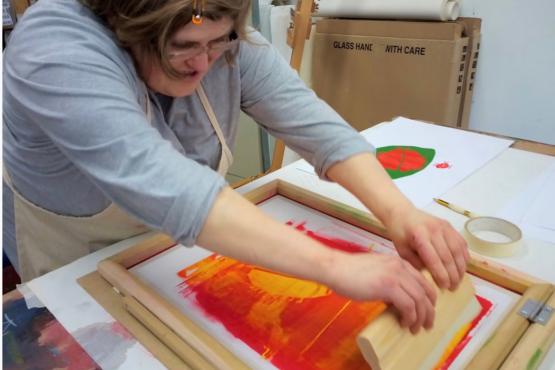 A person screen printing in Shallal Studios. They are using red and orange ink and pulling the squeegee towards them.