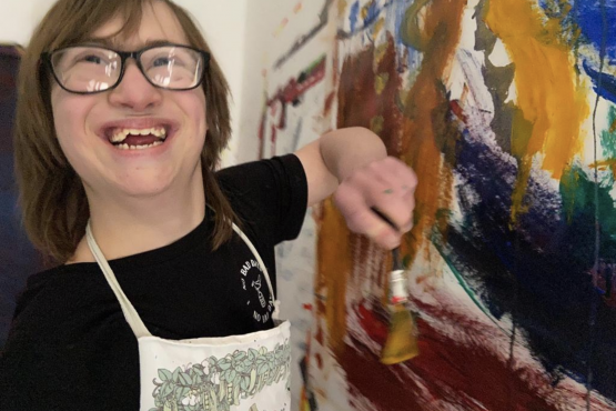 A person smiling at the camera holding a paintbrush. There is a large abstract painting on the wall in front of them.