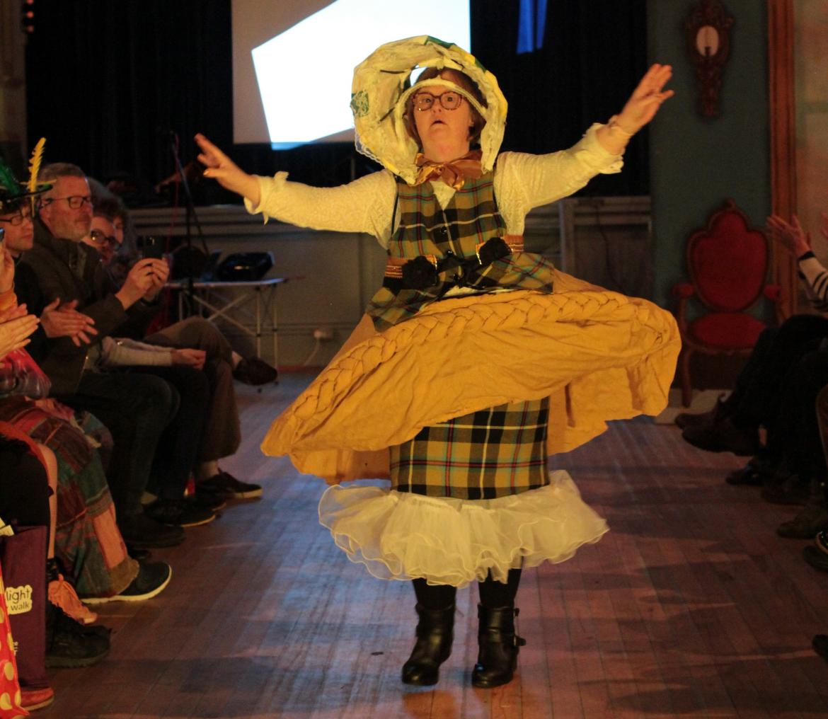 A person is standing at the front of the catwalk in a fashion show. Their arms are raised up elegantly, showing their costume featuring a giant yellow pasty shaped skirt, Cornish tartan waistcoat, white sleeves and decorated bonnet.