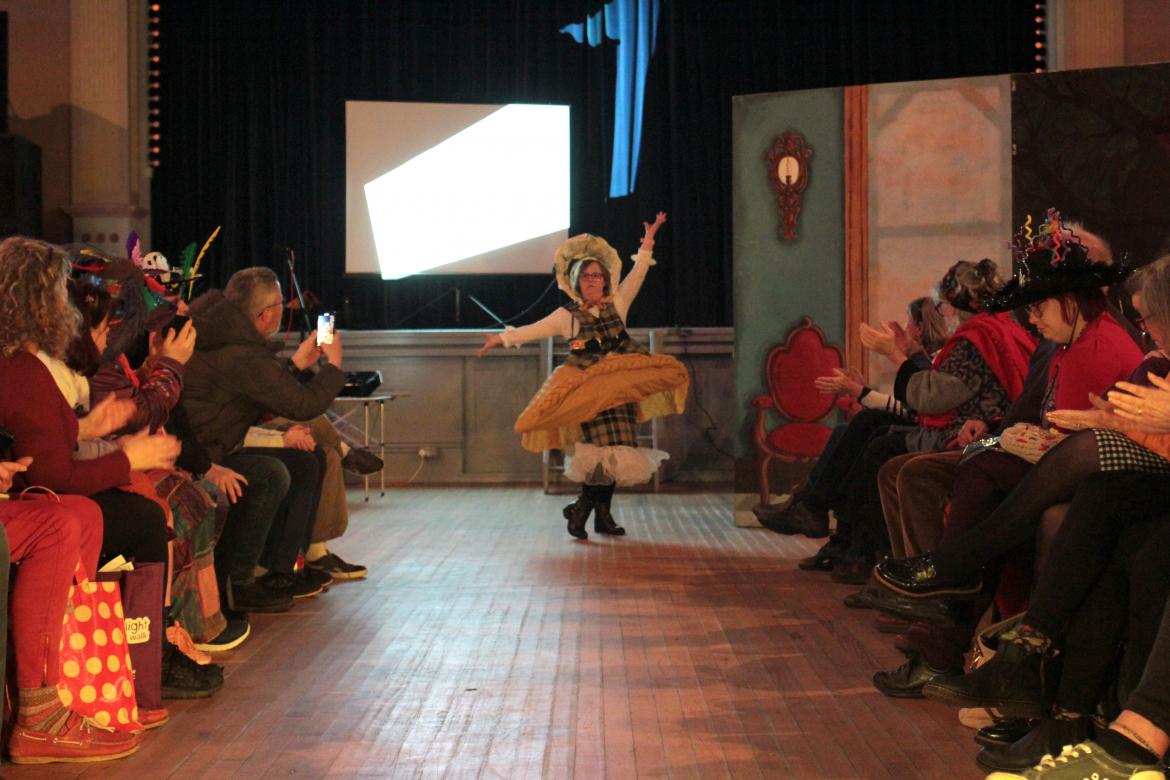 A person is standing at the far end of the catwalk in a fashion show. Their arms are raised up elegantly, showing their costume featuring a giant yellow pasty shaped skirt, Cornish tartan waistcoat, white sleeves and decorated bonnet.