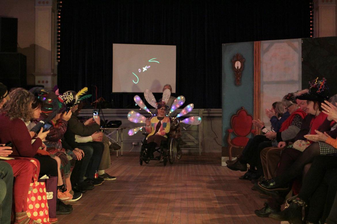 A person is at the far end of the catwalk at a fashion show ready to move forward. The audience  to the left and right are clapping. Their costume includes their wheelchair, adorned with giant white illuminated petals like a peacock showing it's feathers.