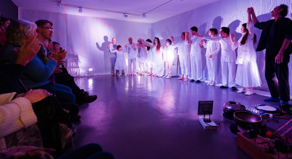 Many dancers preparing to take a bow in a single line on the right of the image. Their hands are joined and in the air and they are bathed in a purplish white light. They are all wearing white except the person who wears black. The audiences is visible on the left.