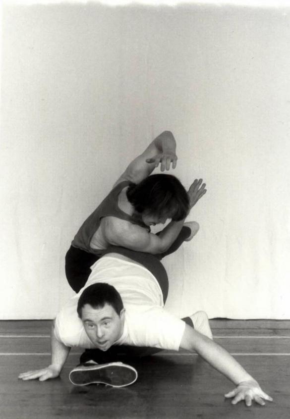 2 people dancing. The image is in black and white. The 2 people are entwined around each other. The first person is on all fours, close to the ground, with one leg in the air and they are looking forwards. The second person is crouched over the other person's legs, they have their body twisted and are holding their arms and hands up so that they frame their head. They are looking towards the floor and one leg is reached out so that it is underneath the stomach of the person on all fours.