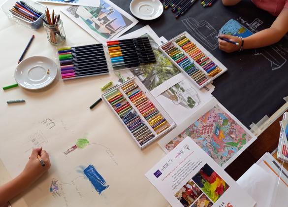 A long table is covered in a long sheet of white paper and a long sheet of black paper side by side. We can see people's arms and hands drawing and colouring on the paper using pastels, felt pens and pencils.