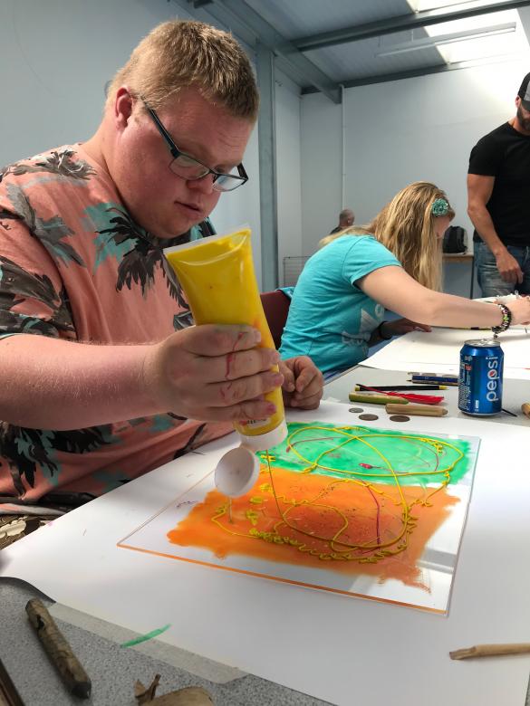 A person sitting at a table monoprinting. They are squeezing a tube of yellow paint onto a piece which is green and orange. There is another person in the background working at the table
