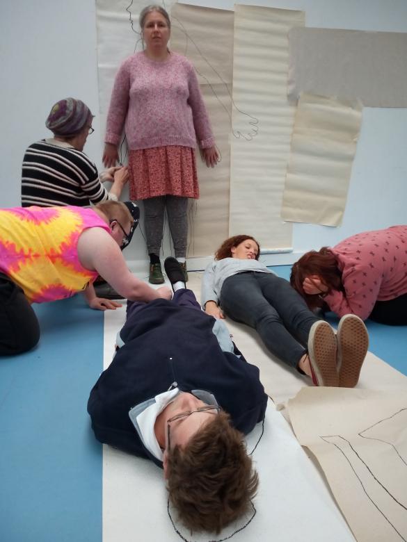 Six people are in a workshop in a room with a blue floor. Two people are lying down whist two other people are sitting on the floor drawing round them on large paper . Another person is standing up and a person is drawing round them onto a large piece of paper fixed to the wall.