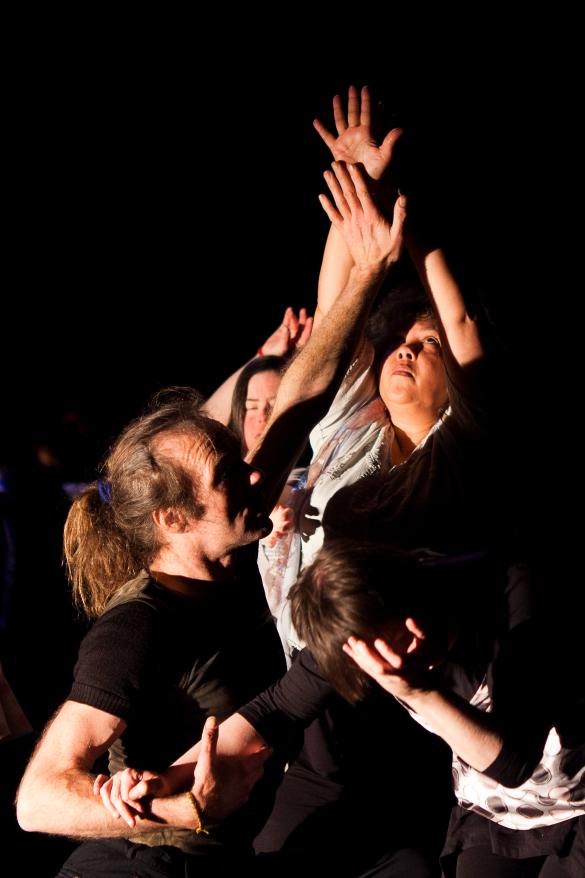 4 people dancing together. The room is dark with their faces and hands illuminated by the light. They are close together and holding hands, which they are reaching upwards.
