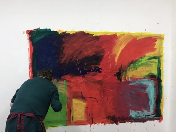 a person with their back turned painting a large colourful abstract painting on the wall. The colours in the painting are red, yellow, dark blue and green