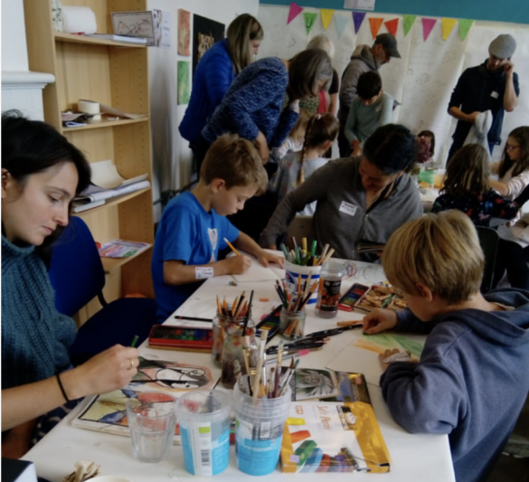 a group of people and children drawing at a table with art materials laid out in sketchbooks. The room is busy. 