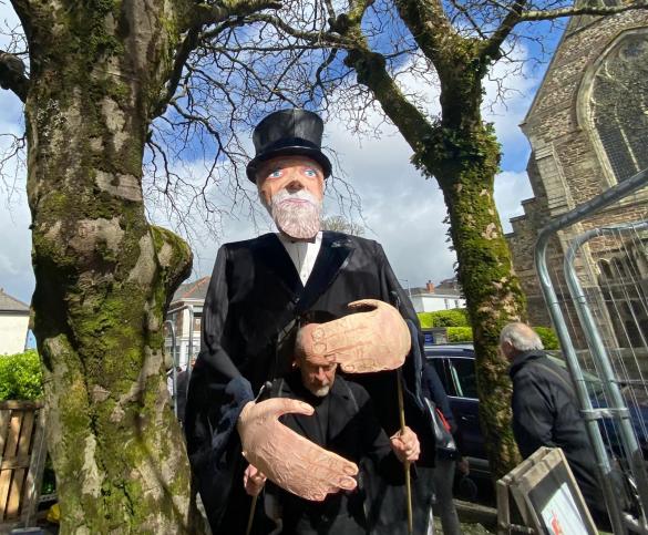 a large puppet with balck clothing and tip hat standing between 2 trees on a street