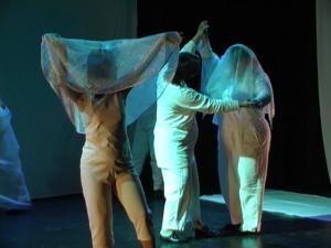 3 people dancing on stage under a blue light. They are wearing white and 2 of them have a shear white scarf over their heads. 