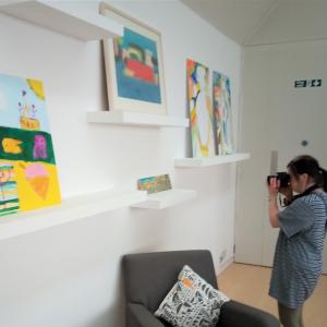 An artist stands in a gallery photographing paintings on the wall
