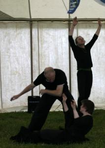 3 people dancing on grass in a tent. They are all wearing black. One person is lying on the ground with their arms raised. The second person is standing bent over the person on the ground and they are swaying their arms. The third person is standing tall at the back raising their arms in the air. 