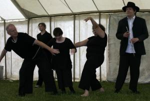 A group of 5 people dancing on grass in a large tent. They are all wearing black. 