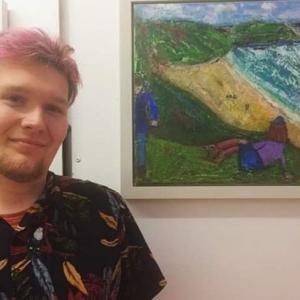 A headshot of Eddie standing by one of his paintings. Eddie is smiling and has pink hair and a black shirt on. 