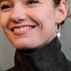A headshot of Sapphire smiling. She is wearing a dark grey high neck jumper and hoop earrings. 