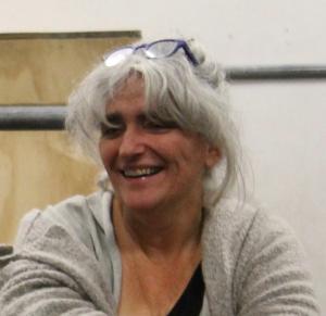 Jo is smiling looking to the left, tanned skin and grey hair, background of  a grey metal bar with wood on the left and white wall on the right. Jo is wearing grey cardigans and a black t shirt.