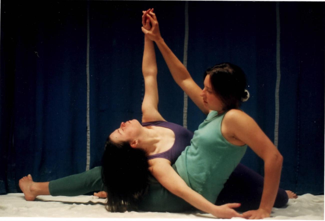 2 people dancing together. They are sitting on the floor facing each other. Their bodies are entwined and they are holding one arm upwards together. The person on the left is leaning backwards