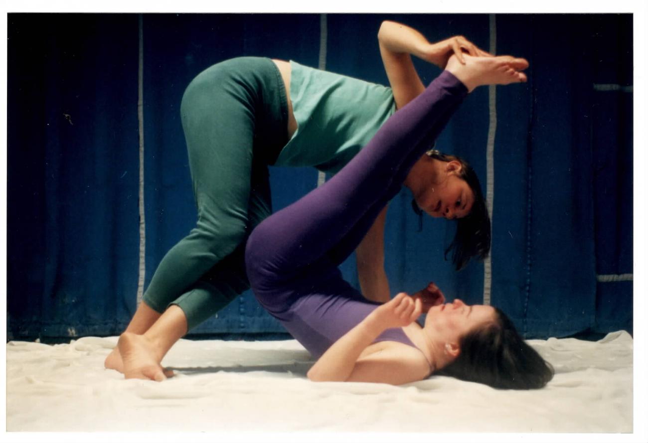 2 people dancing together. One person is lying on the floor on their back with their legs in the air which the other person is holding while standing and leaning over the person lying on the floor.