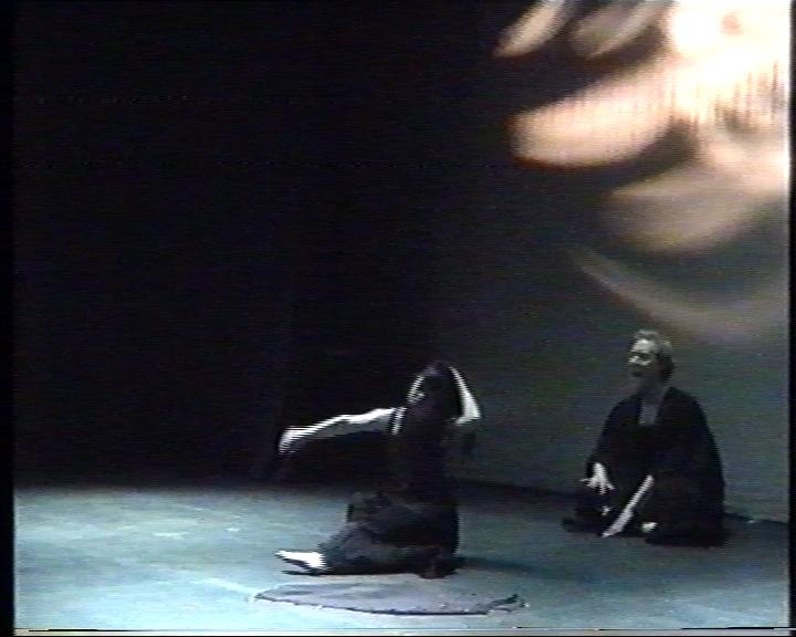 2 people performing on a dark stage. They are sitting on the floor and wearing black. 