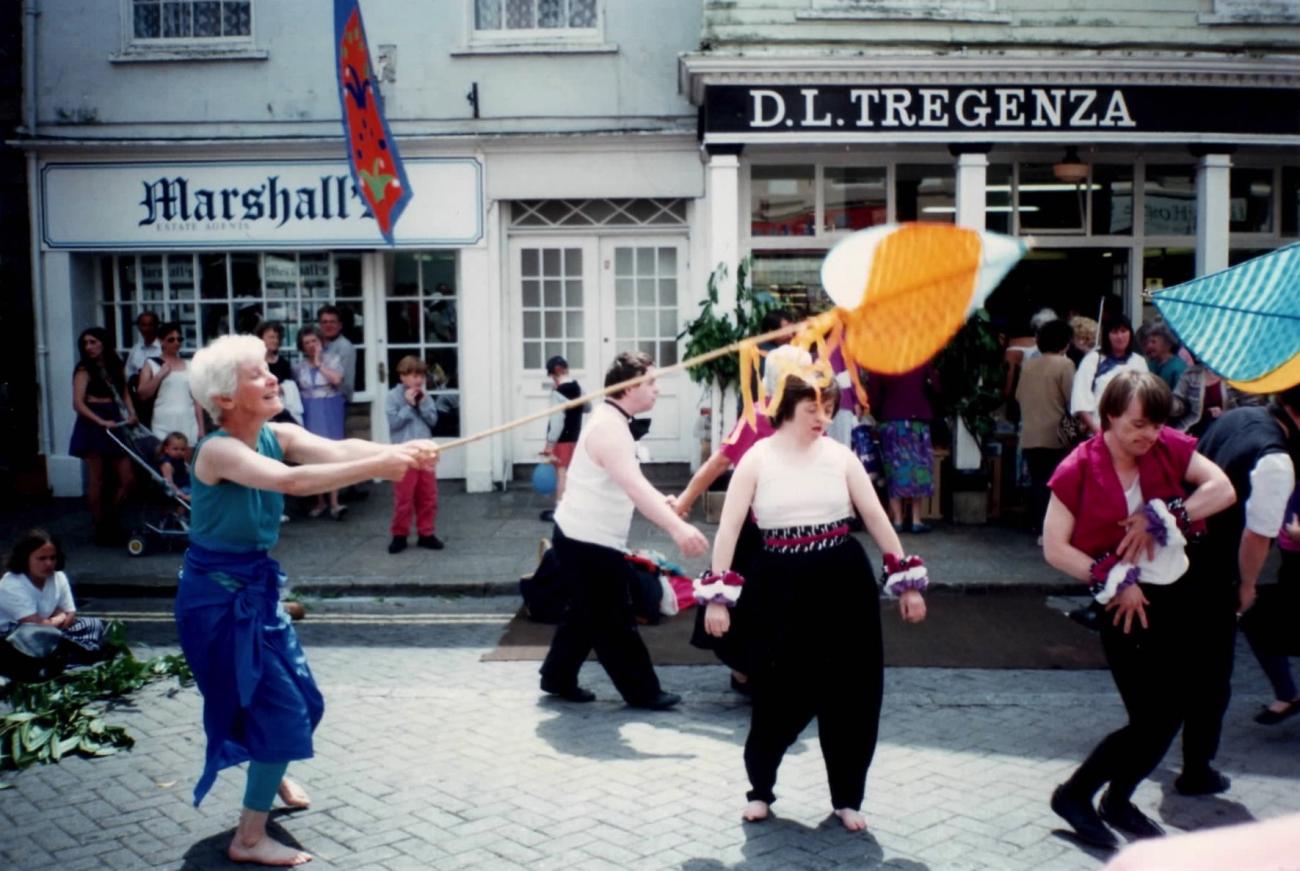 a group of 5 people performing on the street at Mazey Day Parade. One of them is holding a flag. There is a crowd in the background watching. 