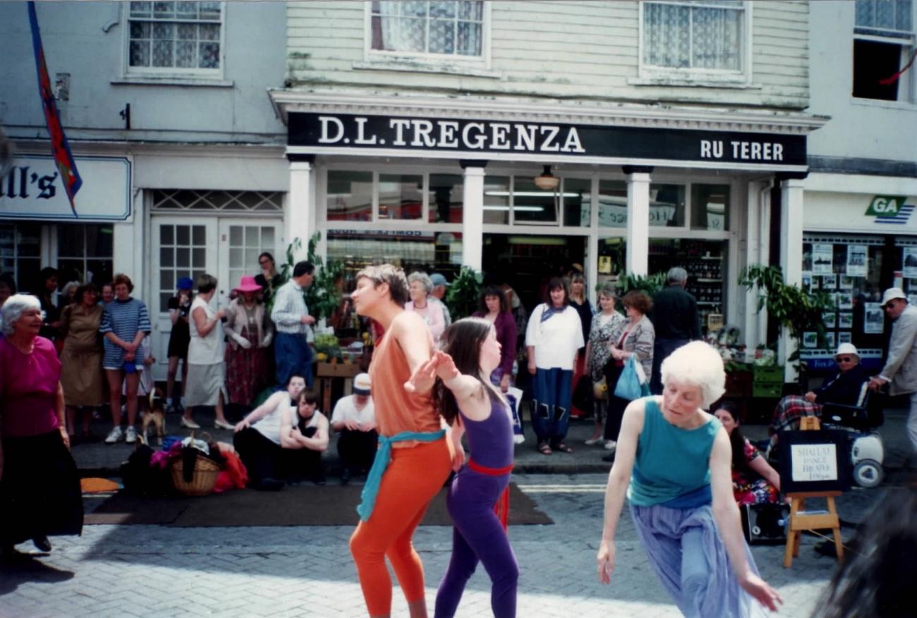 A group of people performing in the street at Mazey Day. They are dancing and their is a crowd around them watching. 