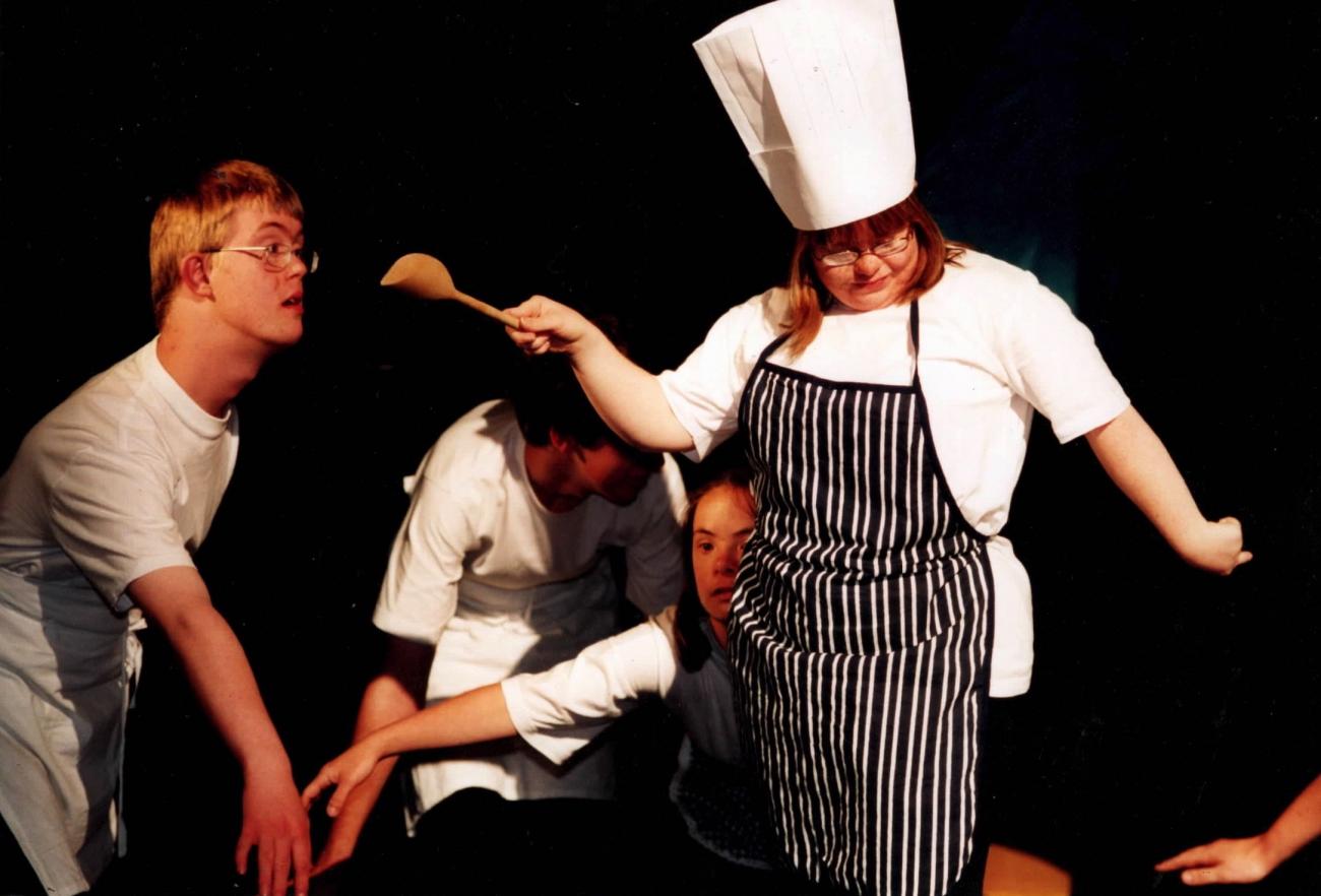 4 people performing on stage. One person at the front is wearing a chefs hat and apron and holding a wooden spoon out. The others are behind this person wearing white and bending forward with their arms reached outwards