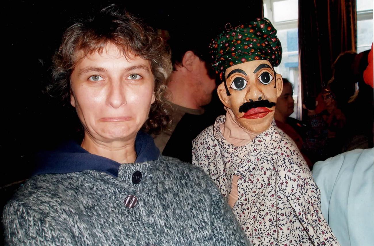 A figure holding a puppet in a crowded room. The figure is looking at the camera and wearing a grey jumper. The puppet has a red and green spotted turban and a floral pink shirt.