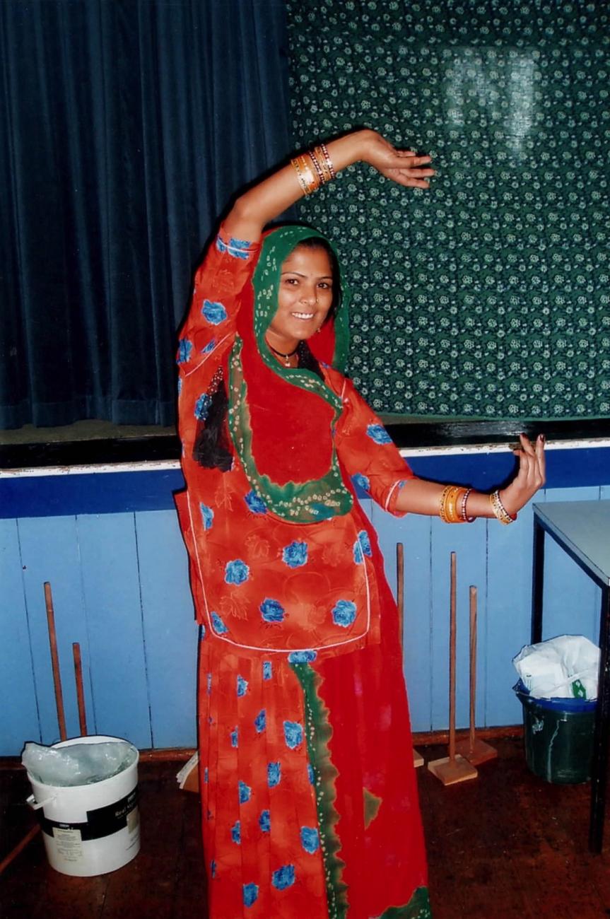 A figure in a red sari with their arms raised above their head and smiling at the camera. 
