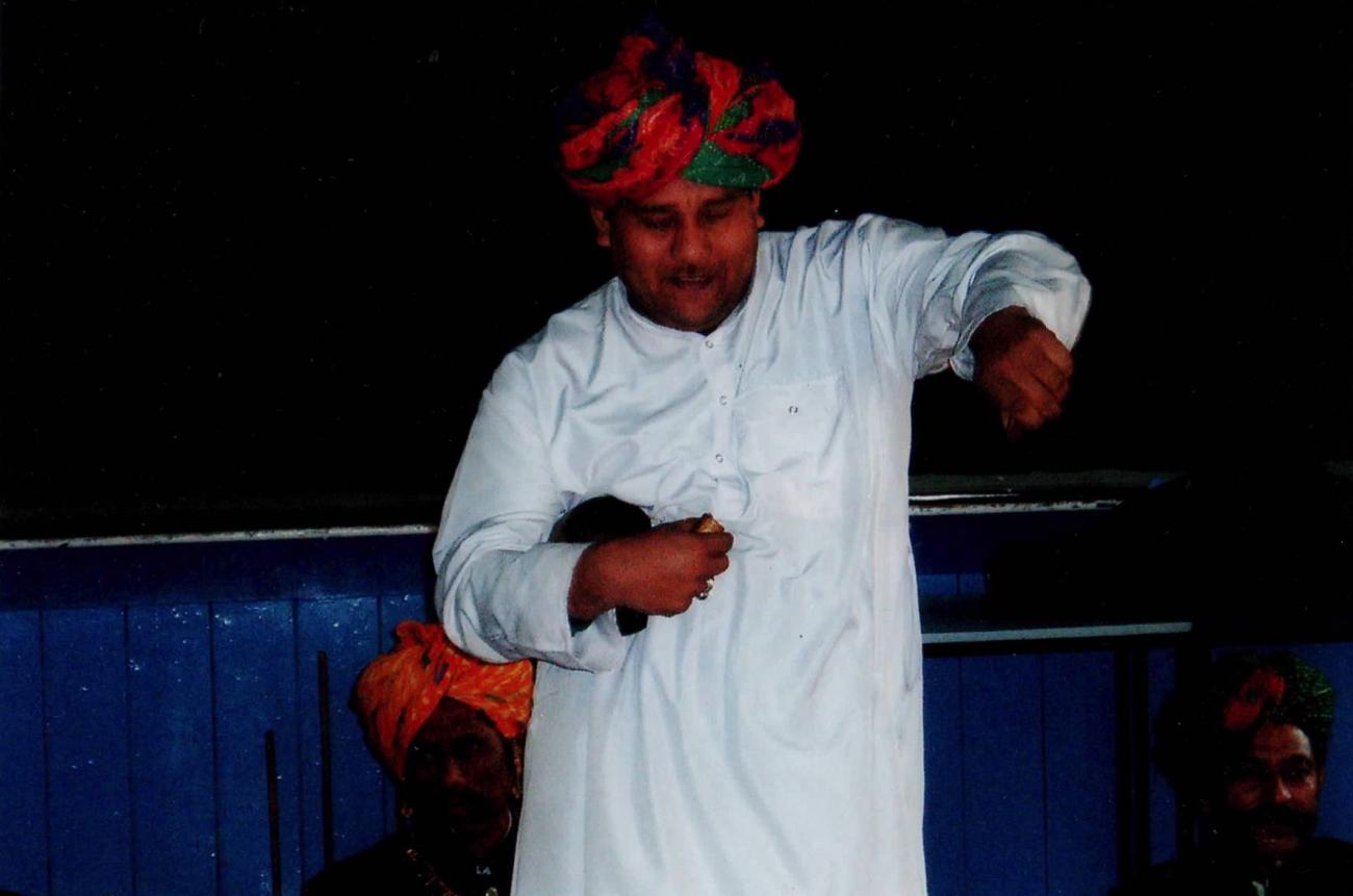 A figure in a long white shirt and a red and green turban dancing. They have their left arm raised and twisted outwards and their right arm is close to their chest holding a small black object. 2 other figures are sitting behind. 