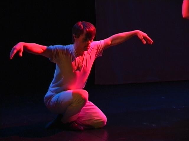 a person dancing on stage. They are wearing all white and are being lit up pink light. They are crouched over with their arms out and up