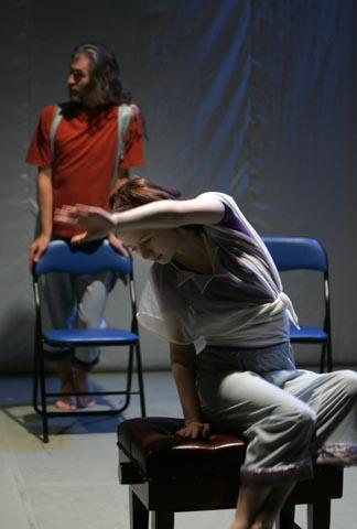 2 people perform in the Mask performance on stage. One in the foreground is seated with one arm raised above their head. The other person is standing leaning on a chair in the background.