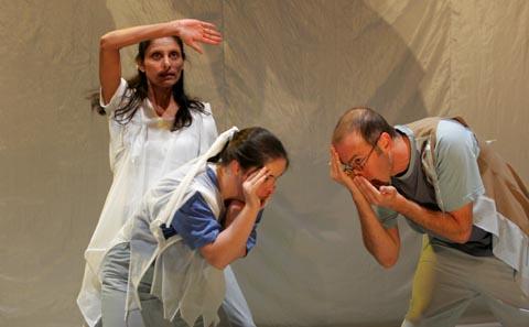 3 people performing on stage. They are wearing white robe like costumes. 2 of them are bent over at the front facing each other and cupping their hands over their face. The third person is in the back with one arm raised up above their head