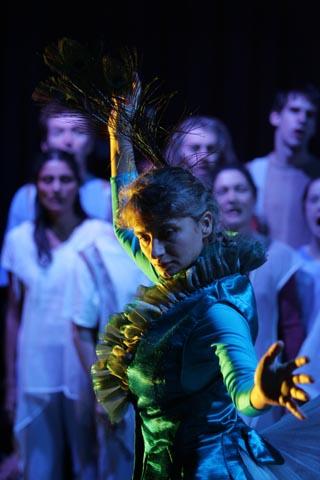 a group of 6 people standing in the background on stage watching a person wearing green with a peacock feather in their hair, dancing in the front