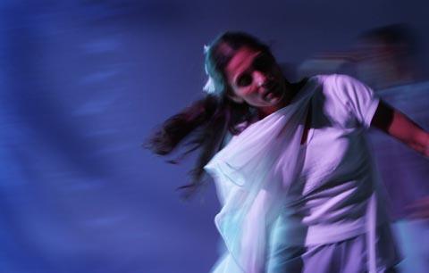 A person with long dark hair dancing in white in a blue lit space 