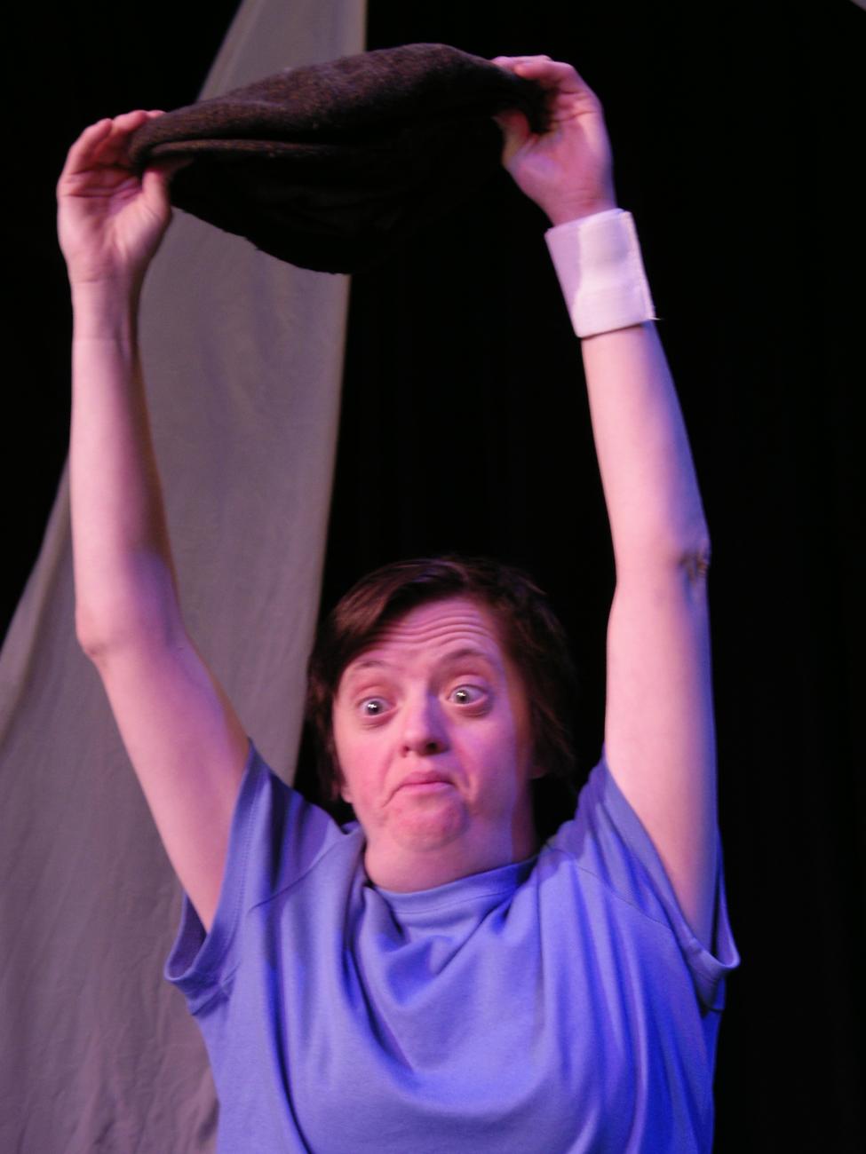A figure performing. They are wearing a blue teeshirt. They have their arms raised above their head and are holding a hat.