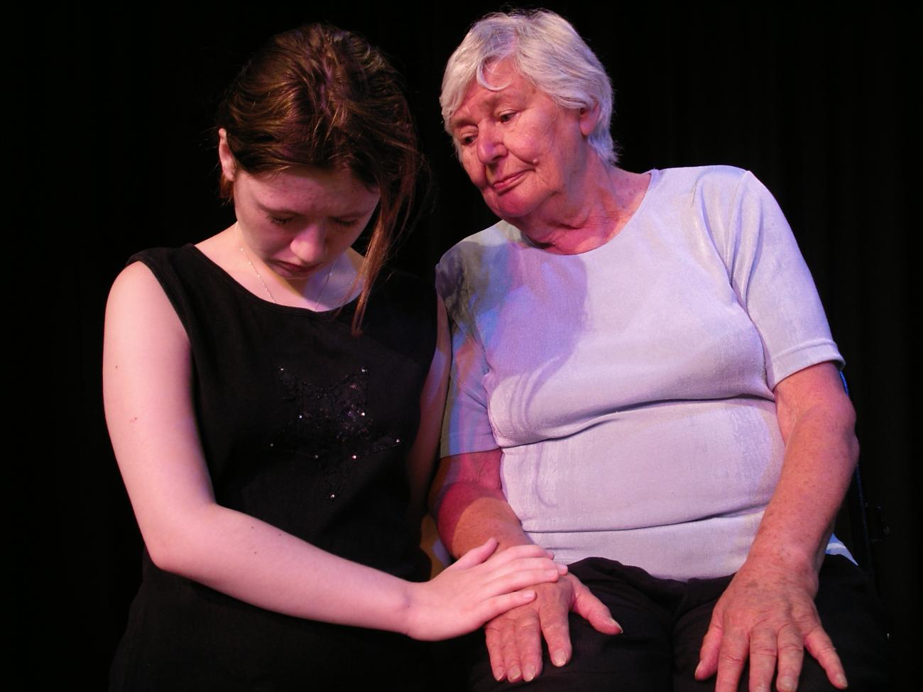 2 people performing. They are sitting close together and holding hands. The person on the left is looking down and the person on the right is looking at the other person. 