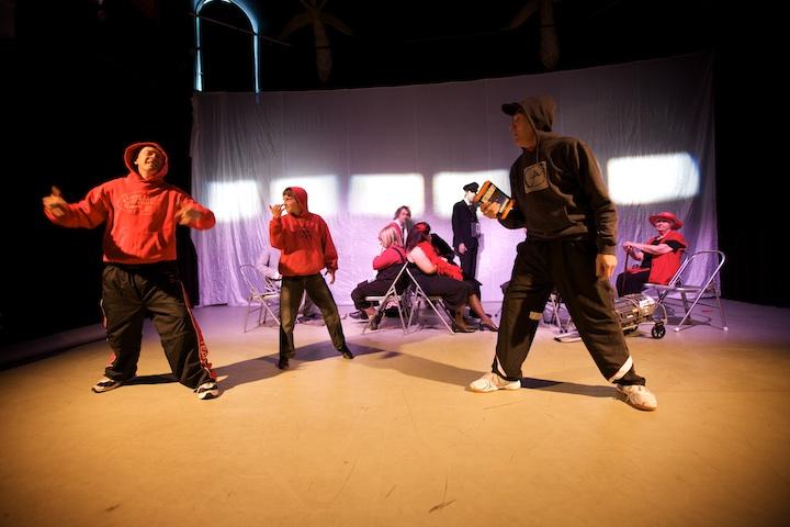 a group of 8 people performing on stage. 3 people are standing in the front wearing red and black hoodies, trainers and black trousers. The other people are sat on chairs in the background.