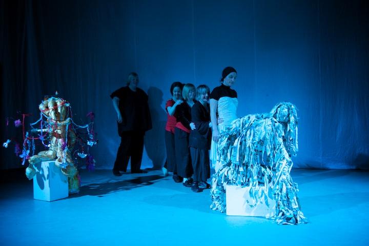 5 people dancing on a blue stage. They are lined up behind each other. There are 2 abstract sculptures on each side of the stage.